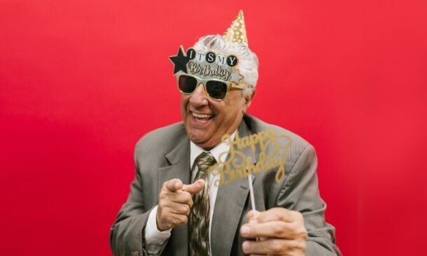 Homme senior en costume, célébrant son anniversaire avec des lunettes festives et un chapeau de fête, souriant et tenant un accessoire Happy Birthday devant un fond rouge.