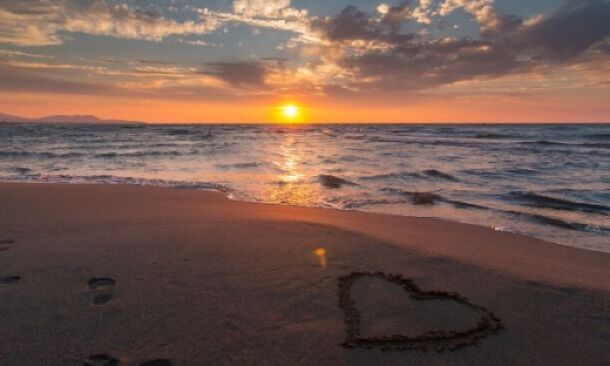 Cœur dessiné sur le sable d'une plage au coucher du soleil, avec les vagues de la mer en arrière-plan et des empreintes de pas à proximité, créant une ambiance romantique et sereine.